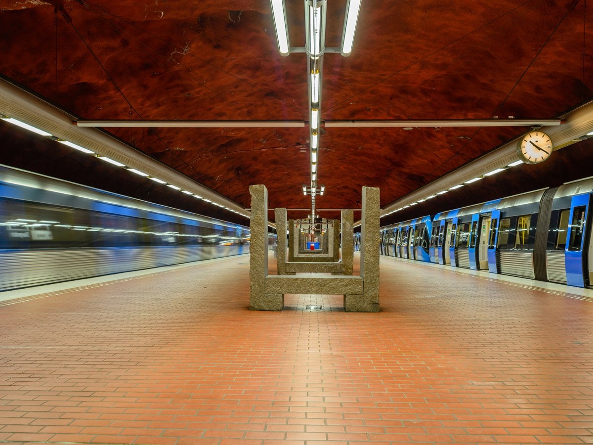 Bild från perrongen på Sparpnäcks tunnelbanestation. Två tåg inne på perrongen, ett på vardera spår, där det ena står still och det andra rullar.