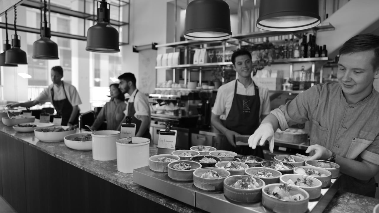 Vy över ett restaurangkök där fem personer jobbar med att tillaga och servera mat