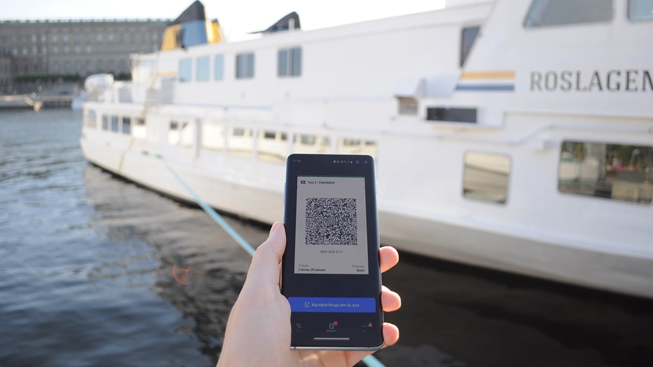 BIld på en hand som håller i en telefon med en QR-kod på skärmen. I Bakgrunden syns slottet och en färja