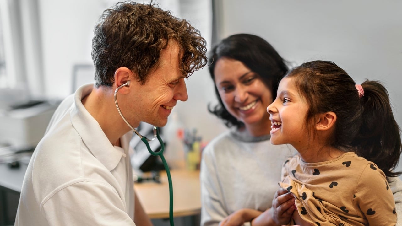 En medarbetare inom vården undersöker ett barn. Barnets mamma syns i bakgrunden.