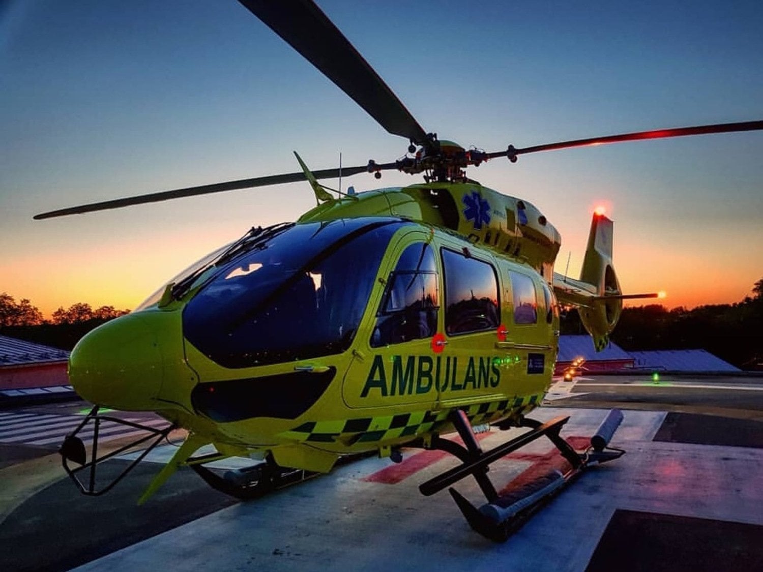 En ambulanshelikopter står och väntar på uppdrag. I bakgrunden ser vi en solnedgång.
