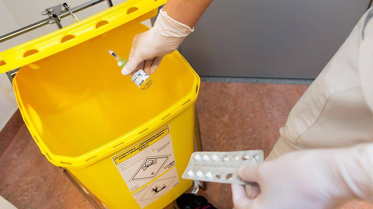 Personer med vita plasthandskar slänger läkemedel i en stor gul återvinningslåda. 