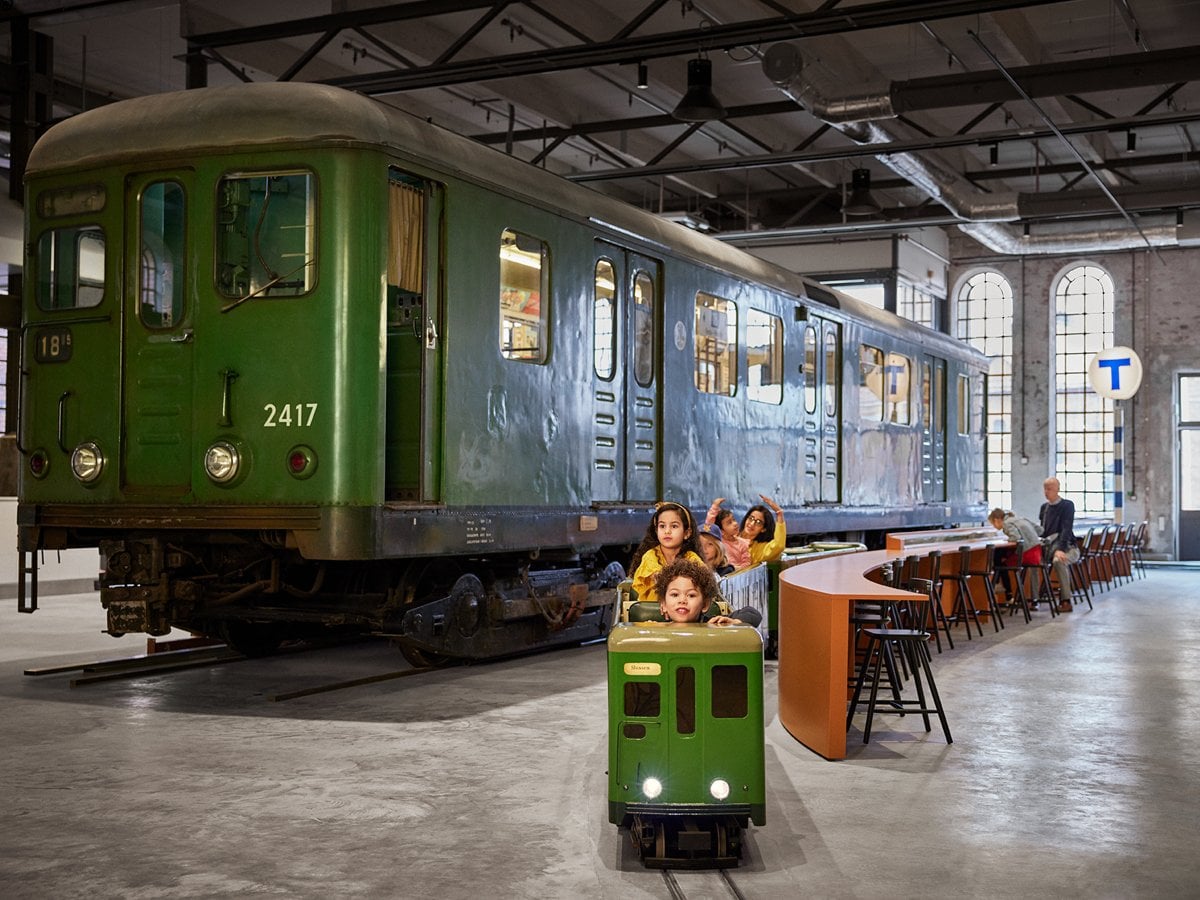 En grupp barn sitter i ett liten tunnelbanevagn, med en stort utställningståg i bakgrunden.