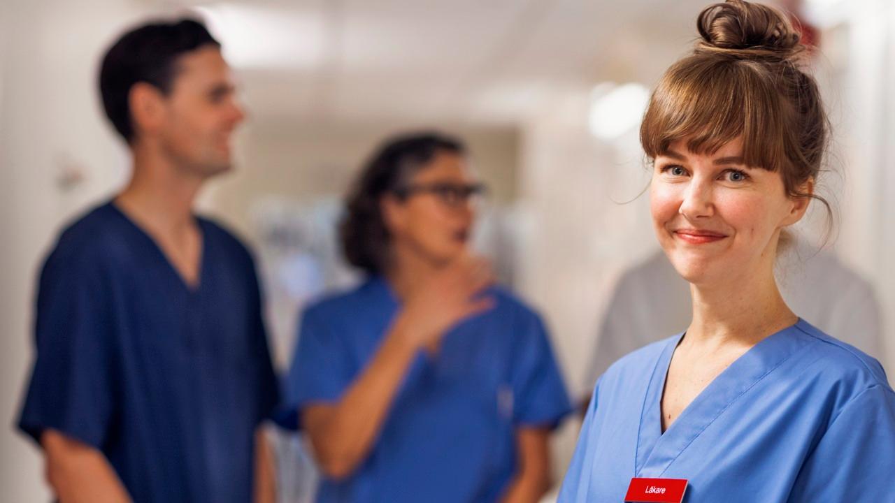 En kvinnlig vårdmedarbetare tittar in i kameran, i bakgrunden står två kollegor