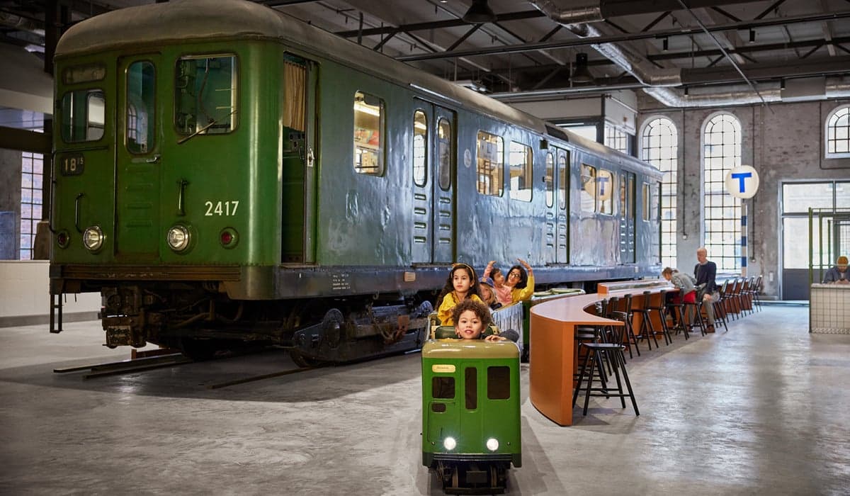En grupp barn sitter i ett liten tunnelbanevagn, med en stort utställningståg i bakgrunden.