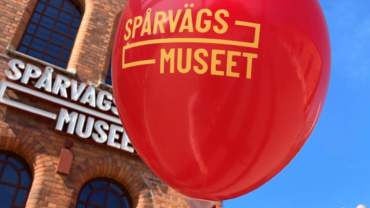 I bakgrunden syns en tegelbyggnad med skylten Spårvägsmuseet. Närmast kameran en röd upplåst ballong med texten Spårvägsmuseet i gult.