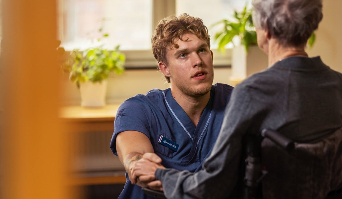 Foto på en vårdpersonal som sitter på huk framför en person i rullstol.