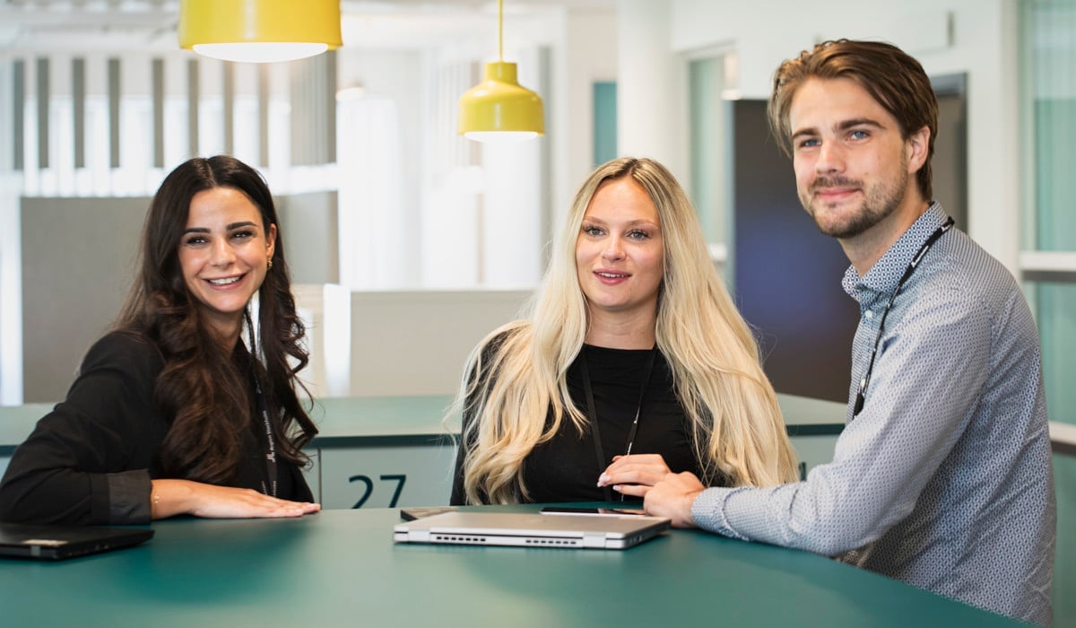 Tre unga kollegor står i ett kontorslandskap vid ett grönt bord och samtalar.