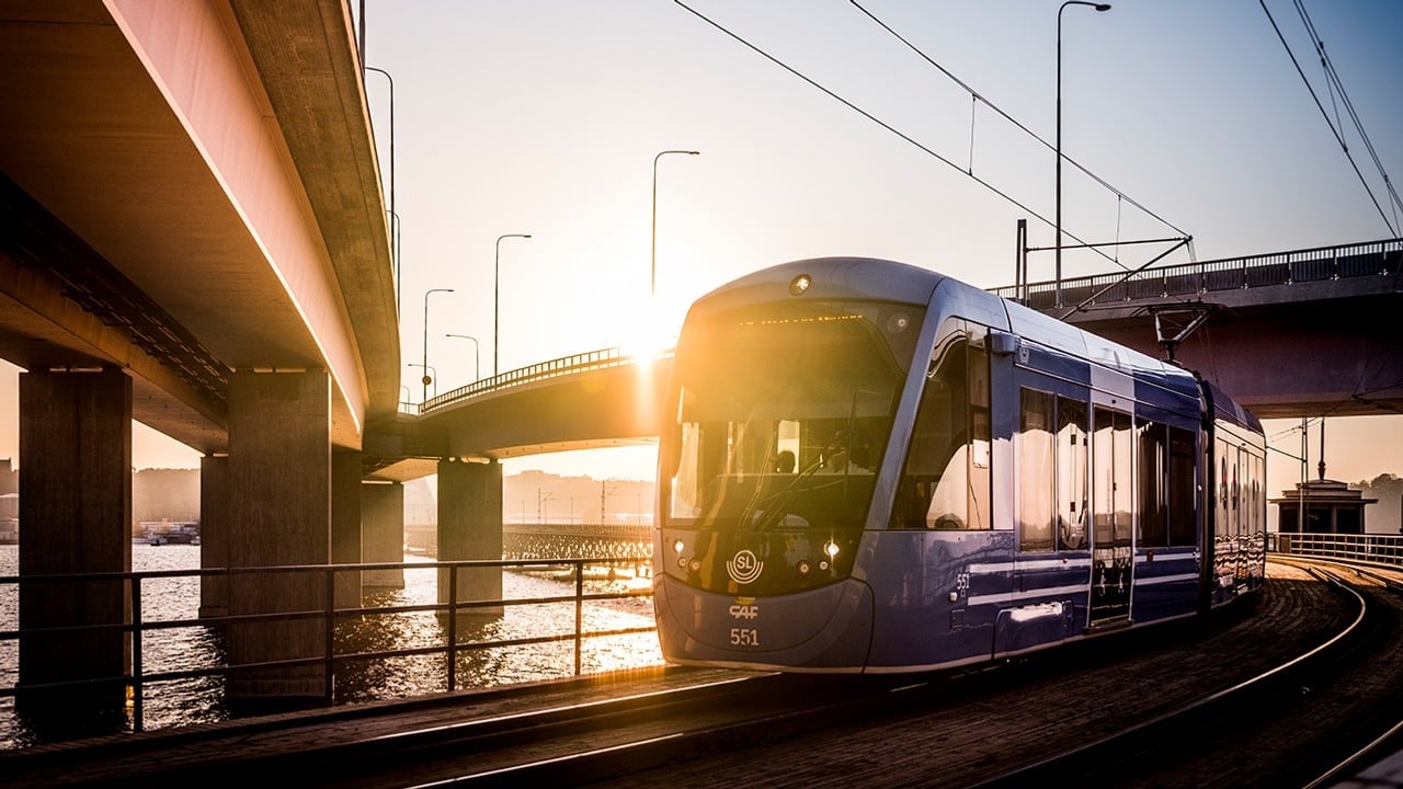 Ett tåg rullar under två broar med stark soluppgång i bakgrunden