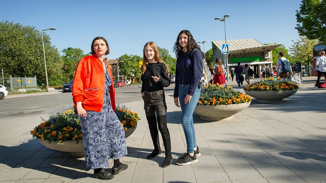 Tre unga kvinnor står och samtalar på ett torg.