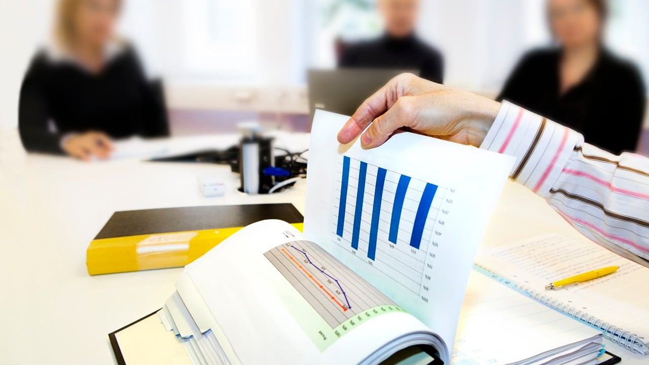 En person håller i ett diagram. På bordet syns flera pärmar, pennor och böcker. I bakgrunden syns tre personer som jobbar vid sina datorer.