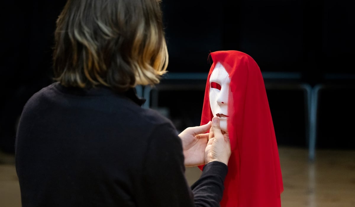 Person står med ryggen mot betraktaren och håller upp en teatermask framför sig.