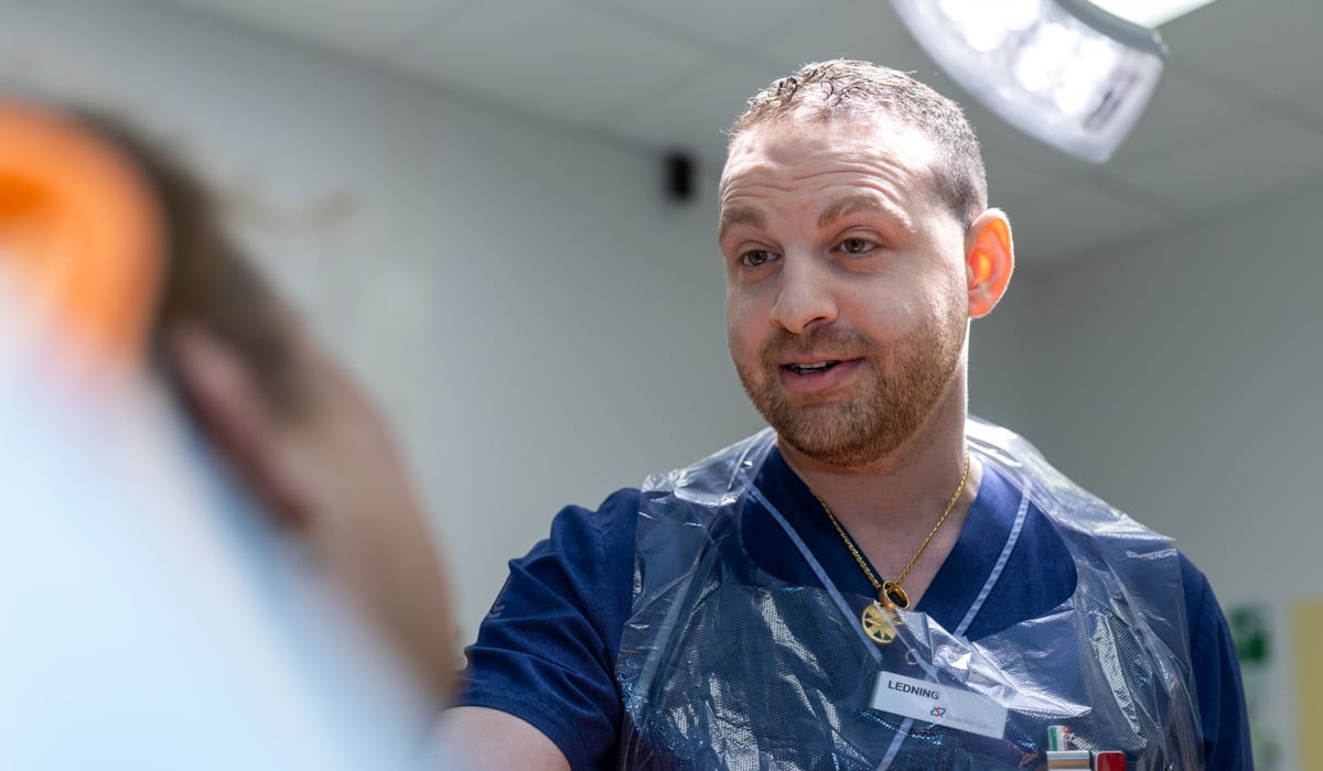 En läkare står framför en patient. Läkaren har sjukhuskläder och plastförkläde.