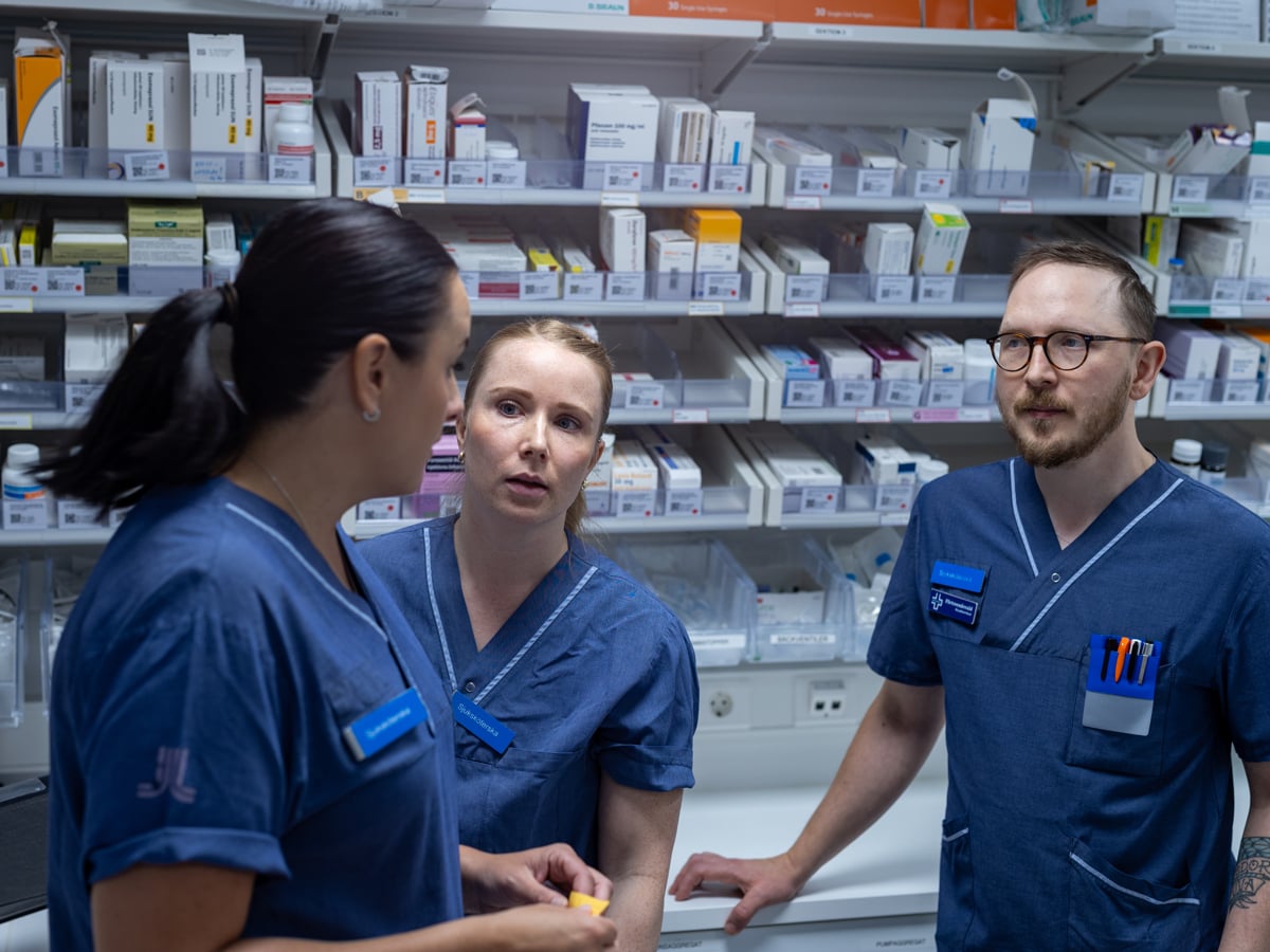 Tre medarbetare i diskussion. De arbetar sannolikt som sjuksköterskor, i samspråk framför vägg av mediciner.
