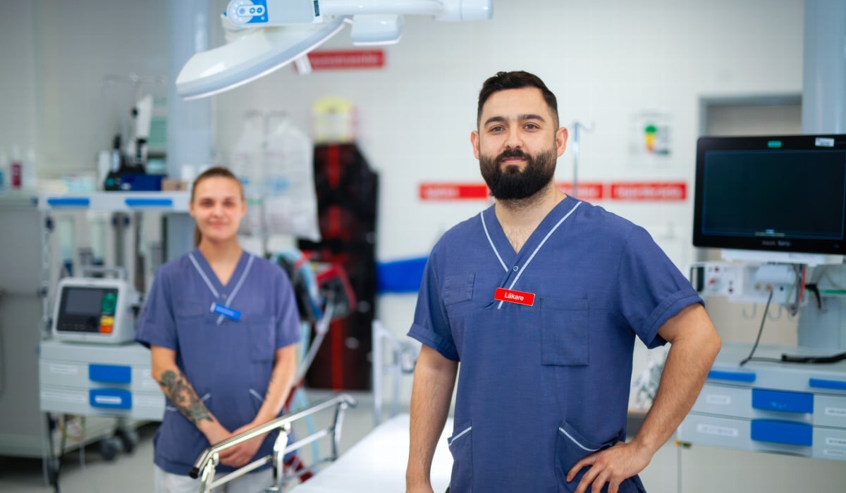 Två sjukvårdspersonal står vid en sjukhussäng och tittar in i kameran.