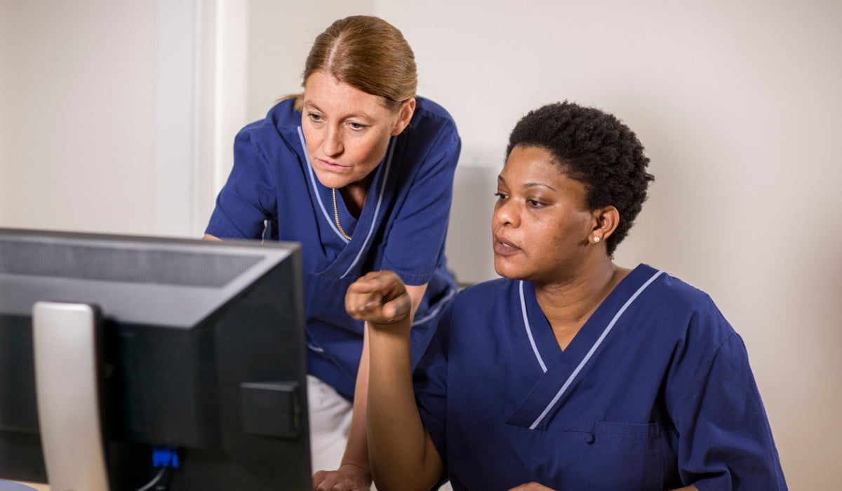 Två kvinnor i sjukhuskläder sitter framför en dator.