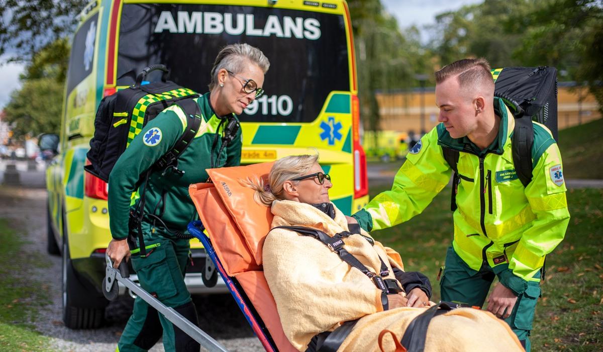Två medarbetare inom ambulanssjukvården i neongröna arbetskläder tar hand om en kvinnlig patient som sitter på en bår.