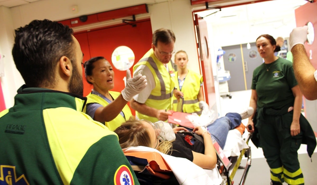 Vårdmedarbetare övar på katastrofmedicin. En kvinna ligger på en bår och runt henne flertalet läkare, sjuksköterskor och annan vårdpersonal i varselvästar.
