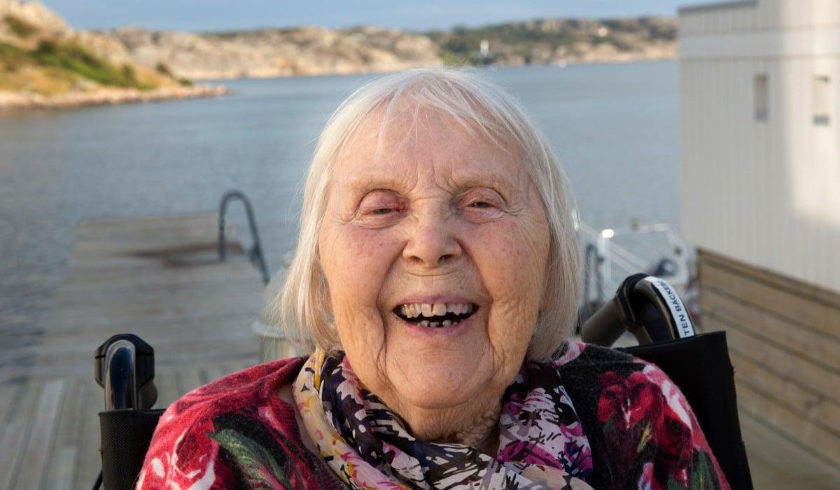 Närbild av en äldre dam som sitter på en brygga och ser skrattande in i kameran, i bakgrunden syns hav och klippr