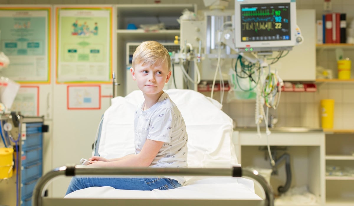 En ung pojke sitter i en sjukhussäng.