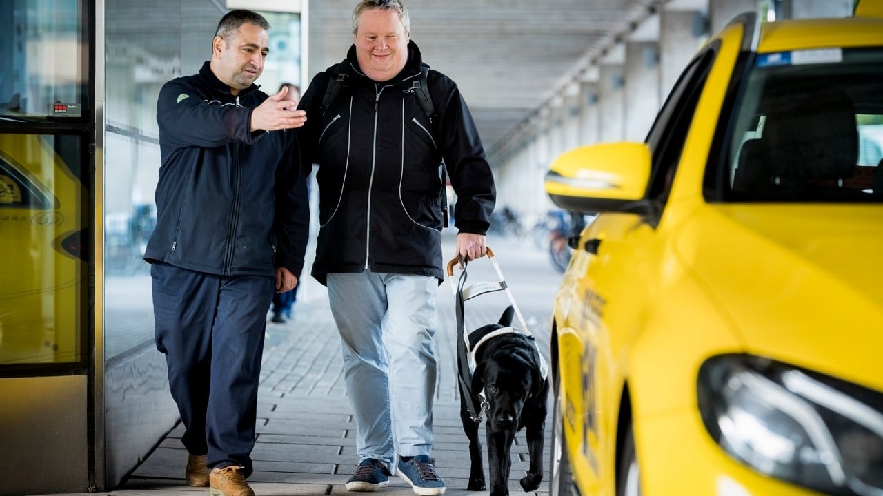 Person med synskada går tillsammans med sin hund. Personen får hjälp att ta sig till färdtjänstbilen.