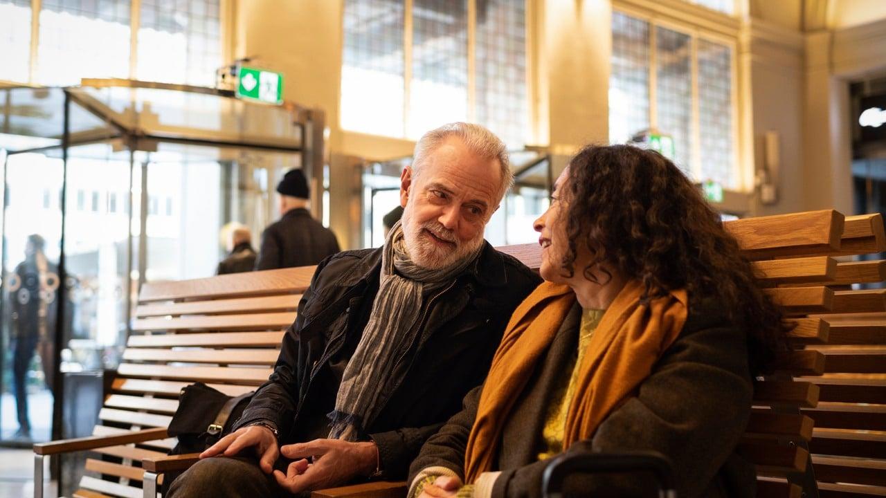 En man och en kvinna sitter på en bänk i en vänthall och pratar med varandra