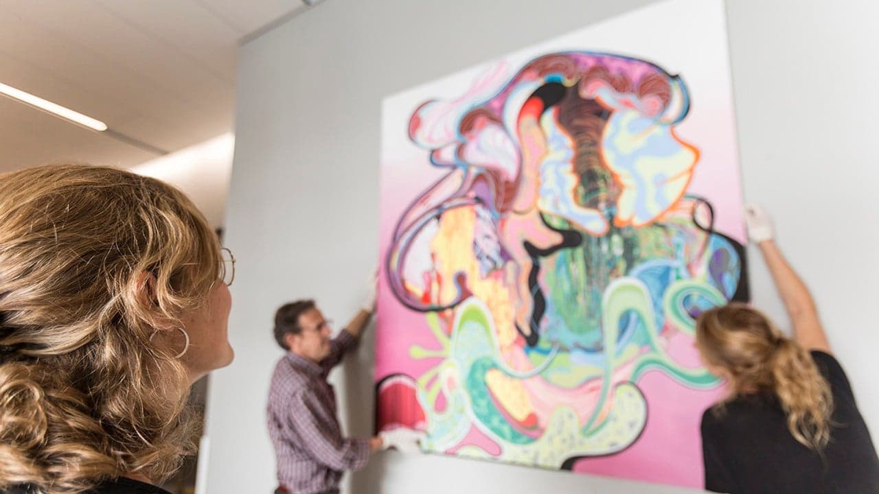 Person står och tittar på upphängning av stor tavla på vägg.