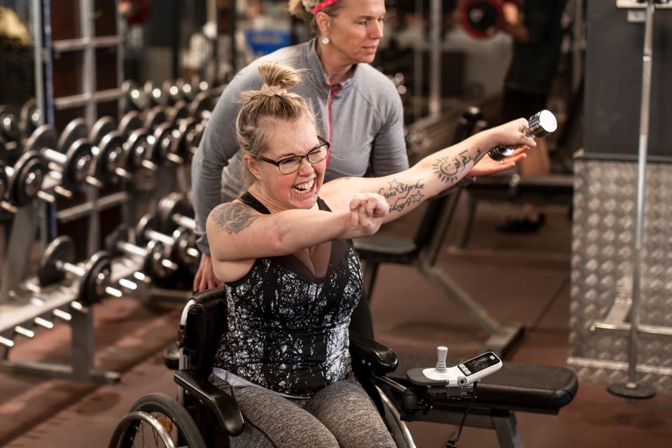 En ung rullstolsburen kvinna tränar med hantlar medan en annan kvinna hejar på