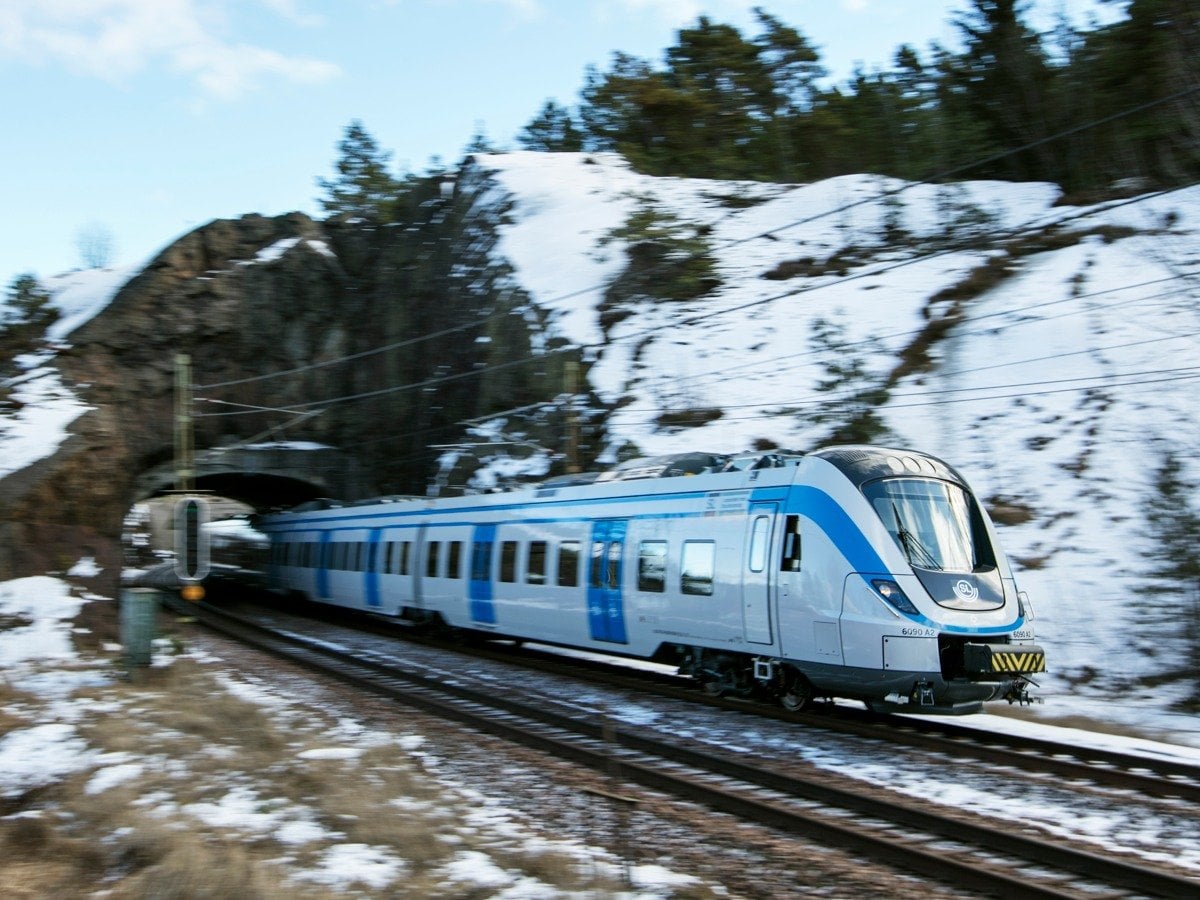 Ett blåvitt pendeltåg åker ut ur en tunnel i hög hastighet. Naturen runtomkring är suddig och bergen är täckta i snö.
