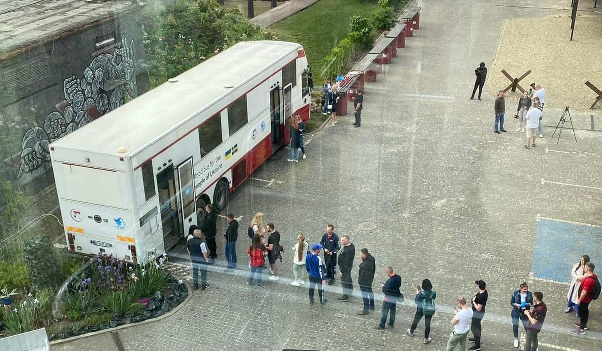 Foto taget uppifrån över ett torg. En blodbuss står parkerad på torget och det är flera personer som köar för att ge blod.