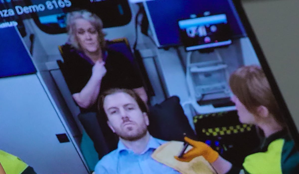 En patient sitter på en bår i en ambulans. Bilden visas på en mobilskärm.