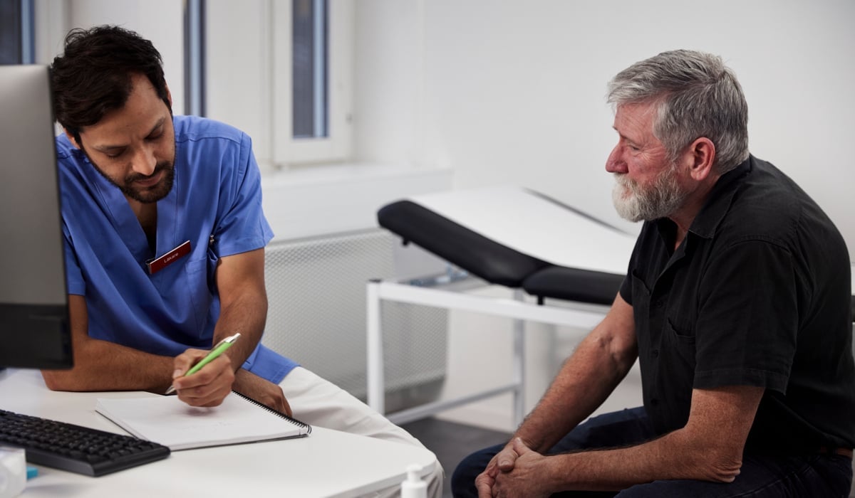Manlig läkare skriver i ett block. Bredvid honom sitter manlig patient.